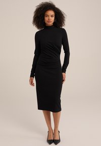 Schwarzes langärmliges Kleid mit taillierter Passform, Rollkragen, gerafftem Taillendetail und mittellangem Saum. Getragen mit schwarzen spitzen High Heels.