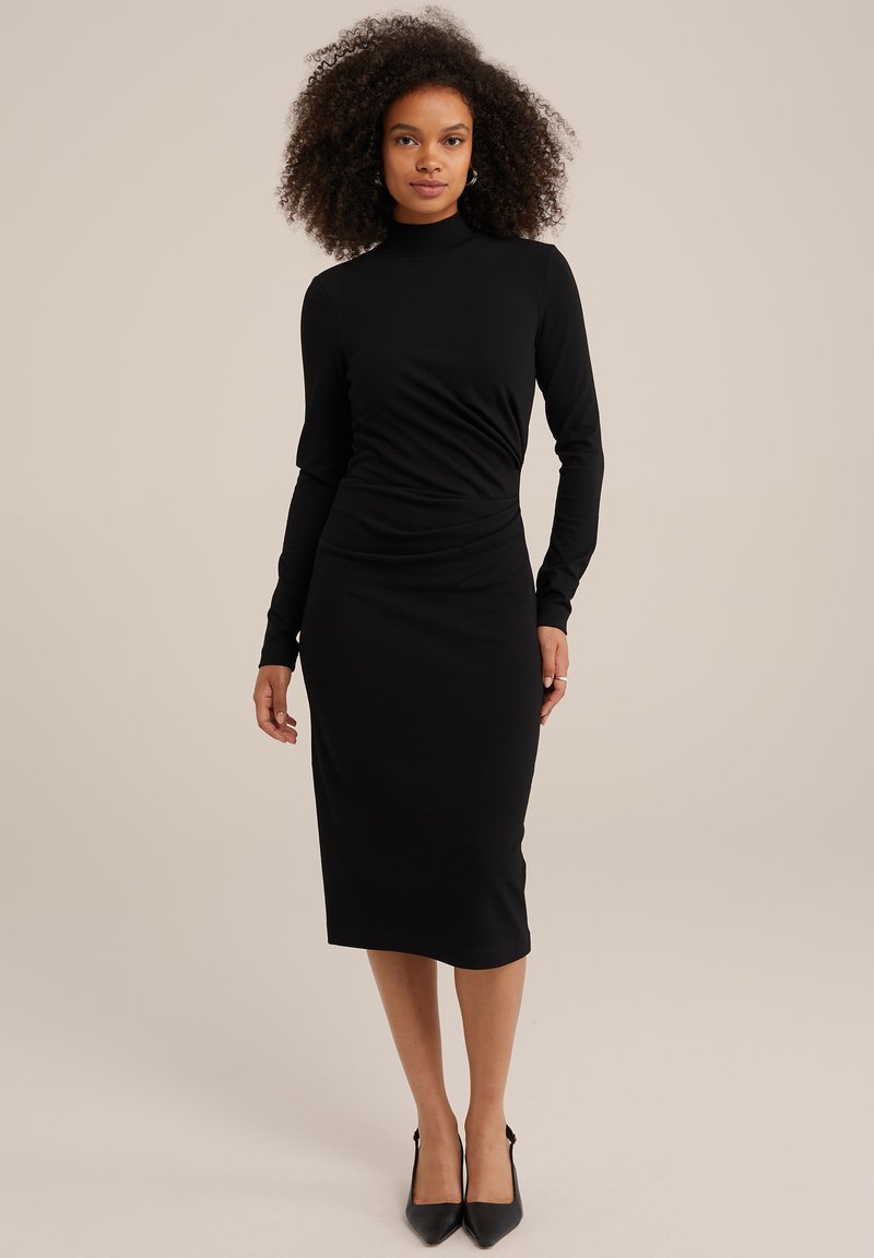 Schwarzes langärmliges Kleid mit taillierter Passform, Rollkragen, gerafftem Taillendetail und mittellangem Saum. Getragen mit schwarzen spitzen High Heels.