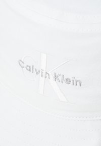 Biely klobúk s širokým okrajom vyrobený z jemnej tkaniny, zdobený tón-v-tóne vyšitým logom "Calvin Klein" a "CK" na prednej strane.