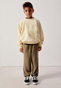 Boy standing facing forward wearing a cream sweatshirt, olive pants, and black-and-white sneakers against a plain background.