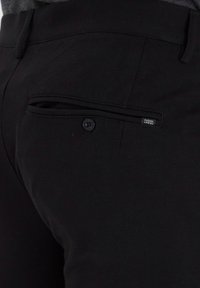 Schwarze lässige Hosen mit einer geknöpften Gesäßtasche, glatter Textur und subtilen Stoffdetails. Das Etikett ist in der Nähe der Taschennäht sichtbar.