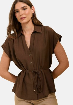 Naine kannab pruuni lühikeste varrukatega nööpidega pluusi, millel on vöö vöökohal, ning beeži värvi pükse, poseerides käed selja taga.