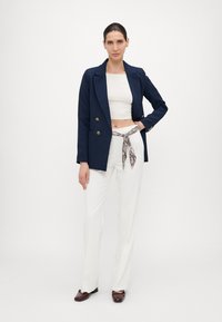 Blazer doppio petto blu navy sopra un crop top bianco, abbinato a pantaloni bianchi a vita alta e una cintura fantasia, completato da mocassini scuri.