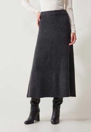 Vrouw draagt een donkergrijze gebreide midirok, een witte top met lange mouwen en zwarte leren laarzen met hak, staand tegen een effen achtergrond.