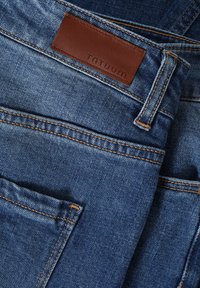 Jeans de mezclilla azules con una etiqueta de cuero que presenta "TATUUM." Las características destacadas incluyen costuras en contraste y múltiples bolsillos.