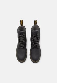 Botine din piele neagră cu șireturi gri, vârfuri căptușite, cusături galbene și etichete în buclă. Se remarcă printr-un design robust și un finisaj texturat.