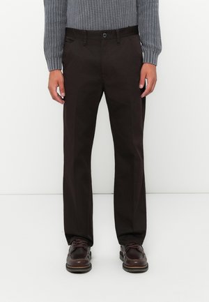 Man wearing dark brown straight-leg pants, dark brown leather shoes, and a gray knitted sweater standing against a white background.