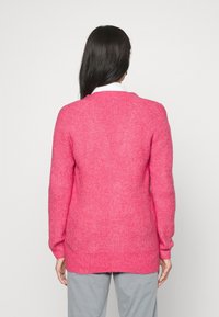 Fuchsia stickad tröja med raglan-design, som har en strukturerad yta och förlängd bakhem, kombinerad med grå byxor.
