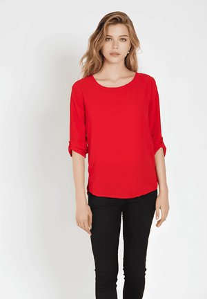 Blusa roja de manga larga con cuello redondo y puños enrollados, hecha de tela lisa. Combinada con pantalones negros ajustados.