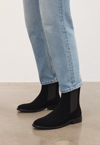 Persona vestida con jeans de mezclilla azul claro y botas Chelsea de ante negras, de pie sobre un suelo de concreto liso.