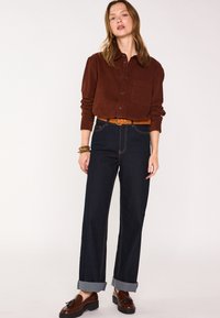 Camicia a bottoni in corda marrone, jeans a gamba larga in denim scuro con risvolti, cintura marrone e mocassini marroni. Design semplice con dettaglio della tasca.