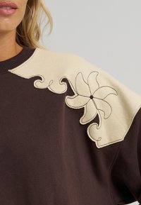 Καφέ φούτερ με κρεμ χρώματος πάνελ στον ώμο, διακοσμημένο με πολύπλοκα κεντήματα λουλουδιών και διακοσμητά ραφές σε κυματιστό σχέδιο.