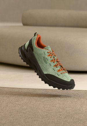 Zapato de senderismo verde claro y negro con cordones naranjas y forro interior, en el aire sobre una alfombra beige y un sofá de fondo.