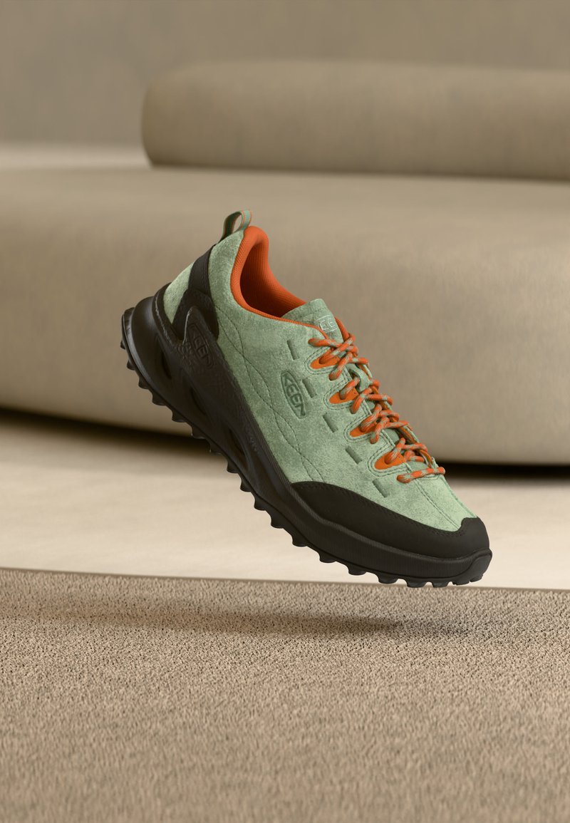 Zapato de senderismo verde claro y negro con cordones naranjas y forro interior, en el aire sobre una alfombra beige y un sofá de fondo.