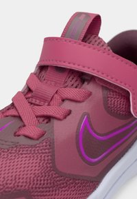 Pantofi Nike de culoare roz, cu un superior texturat, vârful întărit, curea reglabilă și accente violet pe laterale. Au șireturi și o talpă din cauciuc.