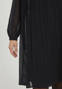 Schwarzes, langärmliges Kleid aus leichtem, transparentem Stoff. Verfügt über geraffte Details am Ausschnitt und elastische Bündchen.