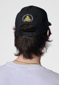 Schwarze Baseballmütze mit gesticktem gelbem Design auf der Rückseite, verstellbarem Riemen und weichem Stoffgefühl.