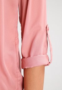 Blouse rose à manches retroussées, en tissu léger. Les poignets sont soigneusement attachés, mettant en valeur une texture lisse.