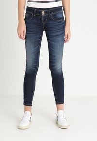 Jeans de mezclilla oscuros, ajustados, con un lavado desteñido, diseño de cinco bolsillos y costuras en contraste. Combinados con zapatillas blancas.