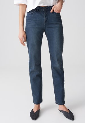 Vrouw met donkerblauwe slim-fit jeans, wit kort topje en zwarte puntige instapschoenen, staand met één hand in de zak.