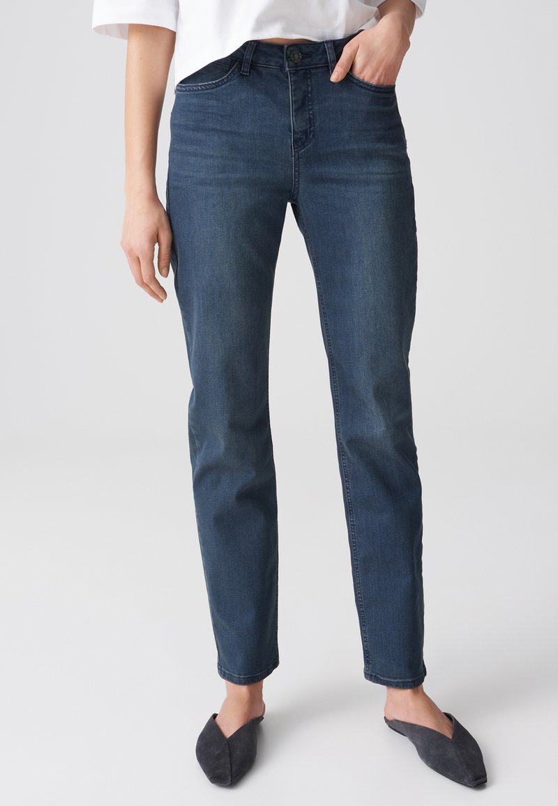 Vrouw met donkerblauwe slim-fit jeans, wit kort topje en zwarte puntige instapschoenen, staand met één hand in de zak.