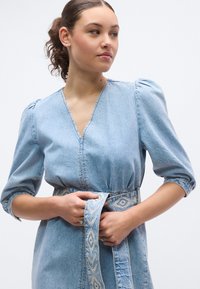 Φόρεμα από ανοιχτό μπλε ντένιμ με φουσκωτά μανίκια, V-λαιμόκοψη, ζωνάκι στη μέση που σφίγγει, και ζώνη με γεωμετρικά σχέδια. Μαλακή υφή.