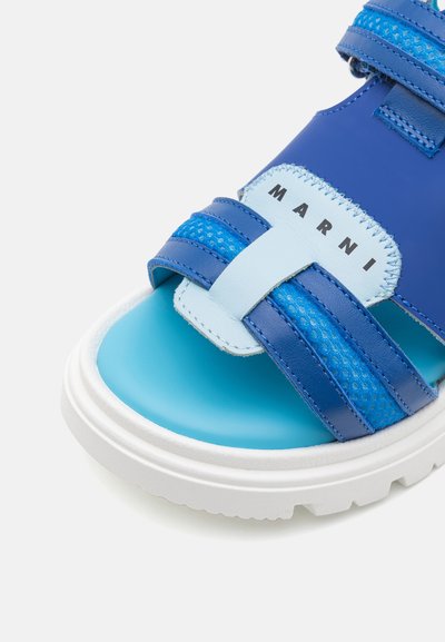 Modra usnjena sandal s mrežastimi detajli, debelim belim podplatom in nastavljivimi trakovi. Ima logotip na strani.