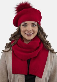 Berretto a maglia rosso con pompon in cima e sciarpa coordinata. Entrambi hanno una texture morbida. La sciarpa si drappeggia liberamente attorno al collo.