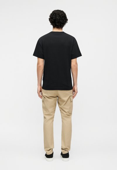 Szembe fordulva áll egy göndör sötét hajú személy, fekete rövid ujjú pólót, bézs színű cargo nadrágot és fekete sportcipőt fehér talppal visel.