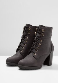 Botines negros con material sintético suave, que presentan ojales en tono dorado, cordones frontales y un tacón bloque grueso.