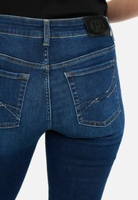 Tmavomodré džínsy z denimu s priliehavým strihom. Medzi ich vlastnosti patrí dva zadné vrecká s dekoratívnym výšivkou z kamienkov a čierna kožená nášivka na páse.