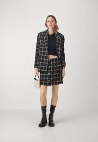 Sort ternetblazer med matchende miniskirt, begge med frynset kant og store knapdetaljer. Båret med skinnende sorte støvler og en lille taske.