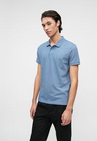 Rövid ujjú világoskék pamut pólóing, gallérral, két gombbal és diszkrét logóval a mellrészen. Fekete nadrággal viselve.