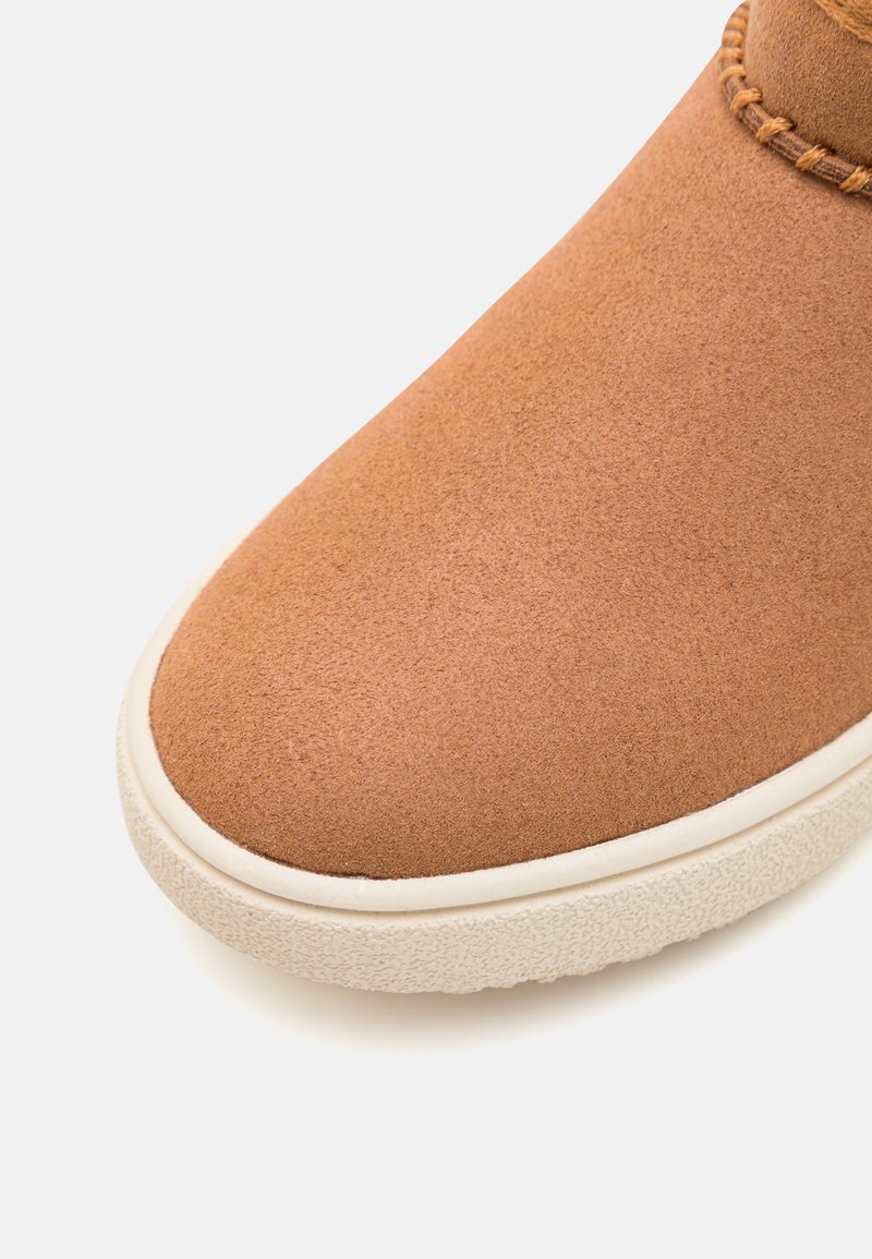 Botín de ante marrón hasta el tobillo con punta redondeada y suela de goma blanca. Cuenta con costuras en el cuello para añadir textura.