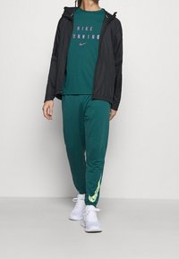 Teal athletic shirt with "NIKE RUNNING" in pink, black lightweight jacket, and green sweatpants with yellow Nike swoosh, paired with white sneakers.