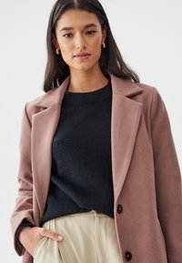 Cappotto in misto lana rosa con colletto a punte, bottoni marroni e texture liscia, indossato sopra un maglione scuro, abbinato a pantaloni color crema.