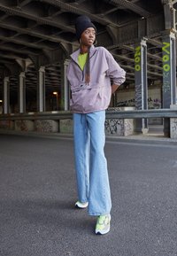 Ljuslila tröja, grön skjorta, lösa ljusblå jeans och vita sneakers med neongrön detaljer. Mot en industriell bakgrund.