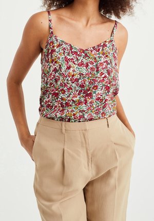 Woman wearing sleeveless floral top with red, white, and green flowers, tucked into high-waisted beige pants with hands in pockets.