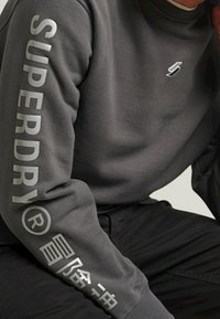 Šedá mikina s ribovaným límcem, na levém rukávu velký reflektující nápis "SUPERDRY" a malé logo na hrudi.