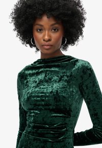 Vestido verde terciopelo con un cuello alto de caída y mangas largas, que presenta una silueta ajustada y un sutil detalle de recogido en la cintura.