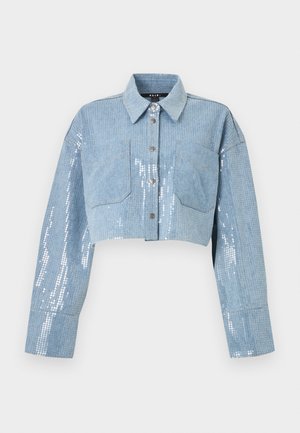 Nainen, jolla on vaaleansininen paljetein koristeltu lyhyt takki ja siihen sopivat leveälahkeiset housut sekä mustat korolliset sandaalit, seisoo valkoisen taustan edessä.