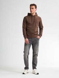 Felpa marrone con cordini, design aderente e piccolo logo sul petto, abbinata a jeans grigi chiari e sneakers bianche con accenti neri.