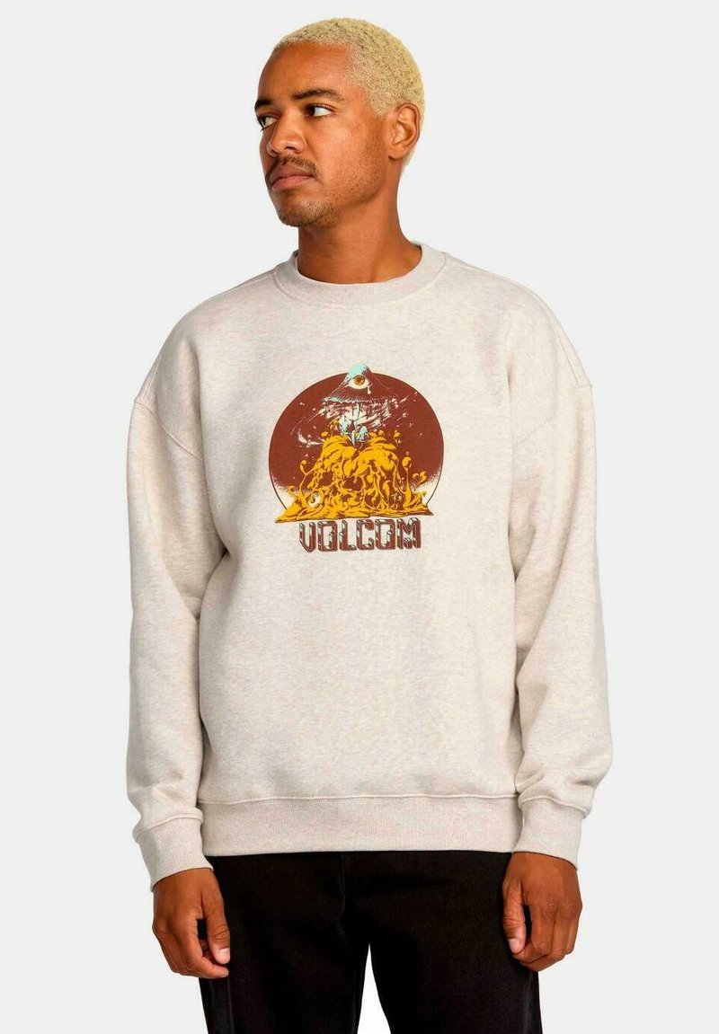 Sudadera beige con un gráfico redondo marrón que presenta un diseño estilizado y el texto "VOLCOM" en la parte inferior. Puños y dobladillo acanalados.