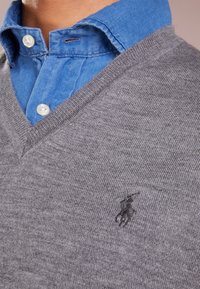 Grå stickad tröja med V-ringning som är lager-på-lager över en blå skjorta med krage. Har en liten svart polo-logotyp på bröstet. Slät textur.