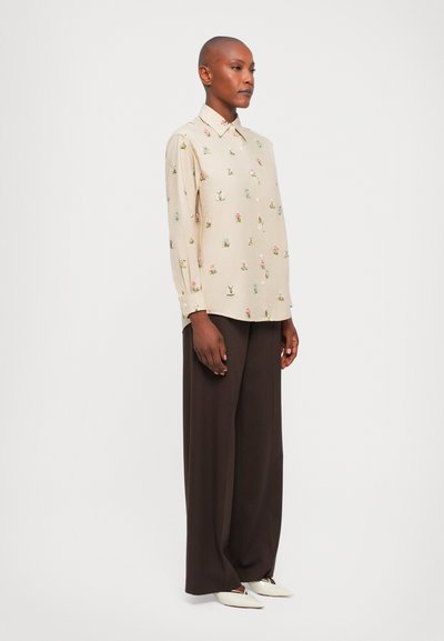 Cămașă bej cu mânecă lungă, decorată cu broderie florală, asociată cu pantaloni largi maro închis și pantofi albi cu vârful ascuțit. Material neted cu guler.