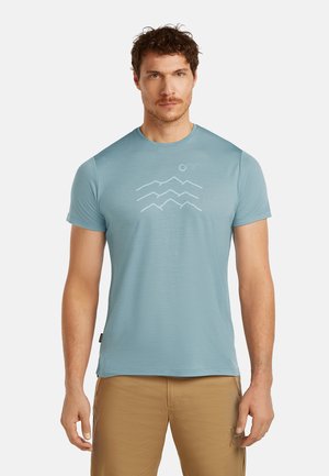 Mann trägt hellblaues T-Shirt mit kurzen Ärmeln und Grafik von Berg und Sonne, dazu beige Hose, steht vor weißem Hintergrund.
