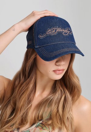 Kobieta z długimi jasnobrązowymi włosami nosząca ciemnoniebieską czapkę Ed Hardy, dotykająca czapki lewą ręką, neutralne tło.