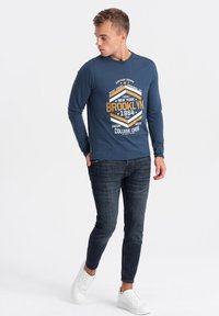 Langärmliges navyblaues Hemd mit einem aufgedruckten Grafikdesign "Brooklyn 1964" in Gelb und Weiß, kombiniert mit dunklen Jeans und weißen Sneakers.