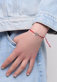 Dłoń z kwiatowym wzorem na paznokciach, nosząca czerwoną bransoletkę ze serduszkiem, spoczywa w kieszeni jasnoniebieskiej kurtki dżinsowej.