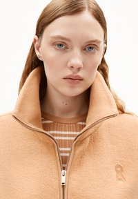 Perzikkleurige fleece zip-up jacket met een hoge kraag en een geborduurd accent. Gedragen over een gestreepte beige trui met een ribbelstructuur.